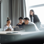 A group of four people in a modern office, focused on laptops and a smartphone