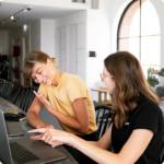 Two women laugh and collaborate at a desk with laptops, in a bright, modern office space
