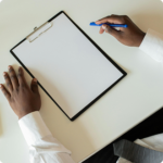 A person in a white shirt holds a blue pen above an empty clipboard on a white desk