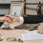 Two people shake hands across a wooden table with a laptop